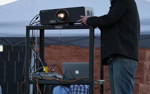 Gregory Crosby wearing a cap and jacket adjusts a projector during an outdoor film festival