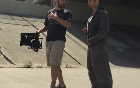 Gregory R.R. Crosby, wearing a baseball cap, t-shirt, and shorts, directs Artur Lago-V who is dressed in a gray hooded sweatshirt during a short film shoot. They stand in a cement ravine with an overpass overhead.