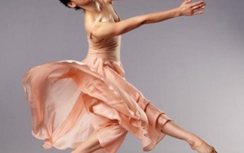 Brenna Monroe-Cook dressed in a flowing peach dress, points her bare foot and reaches out with hands against grey background.