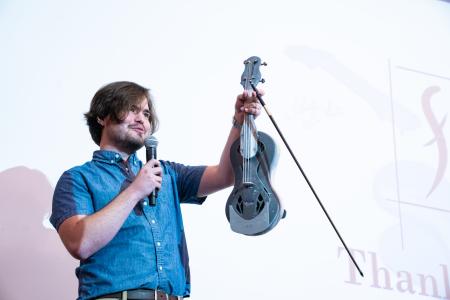 person holding up violin prototype as he speaks into mic