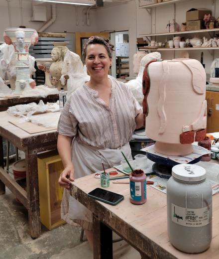 Artist Amy Bessone - white woman in loose shirt and work apron smiles at camera with hand on her partially completed ceramic work