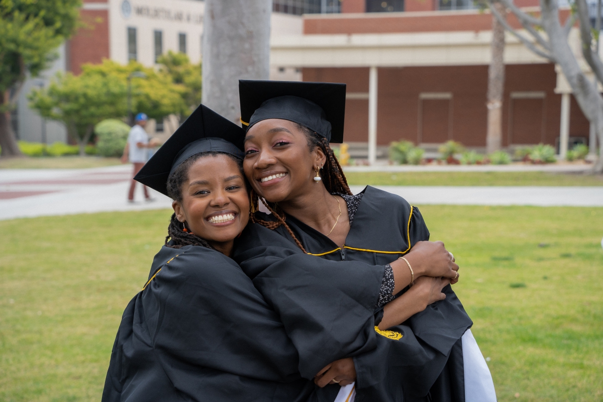 Two students stand hugging and smiling adorned in grad attire.