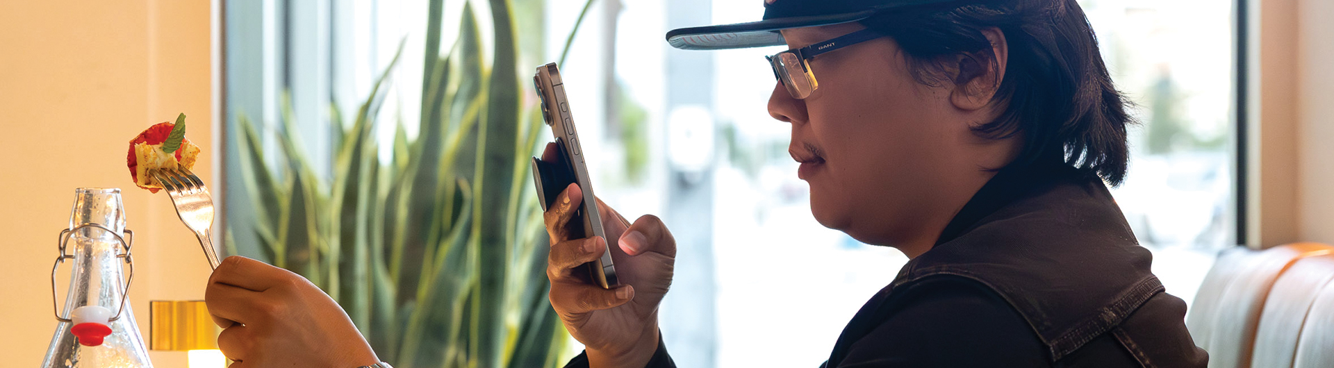 Person sits at a table holding a phone up to photograph a forkful of food.