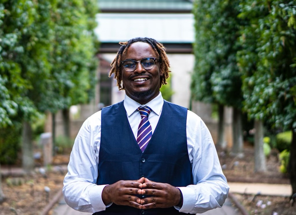 Professor Ky Martin dressed in vest and tie standing outside with trees on either side