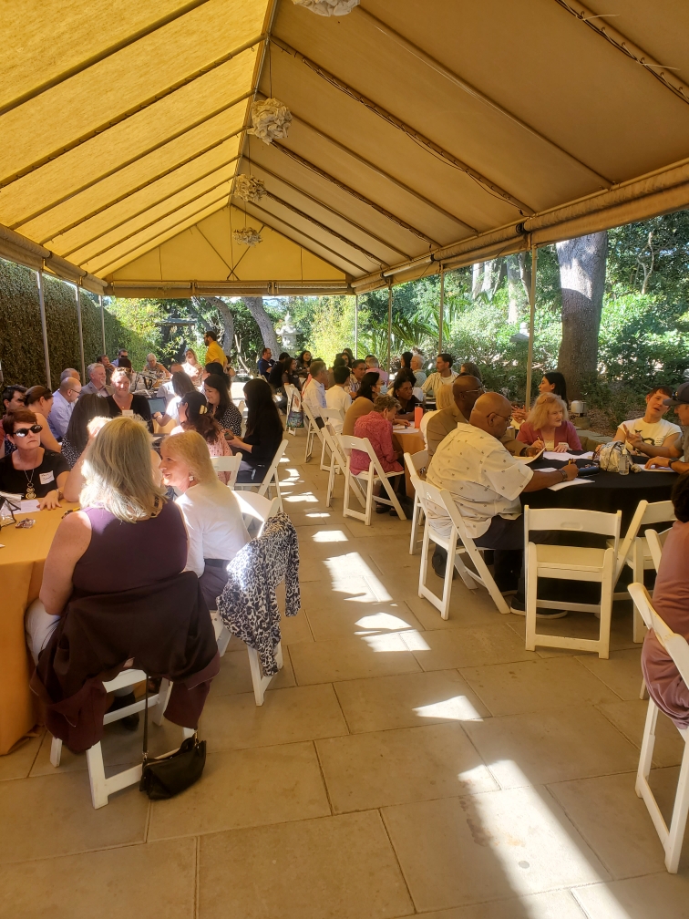 A large group of people sitting at round tables underneath a canopy, talking with one another