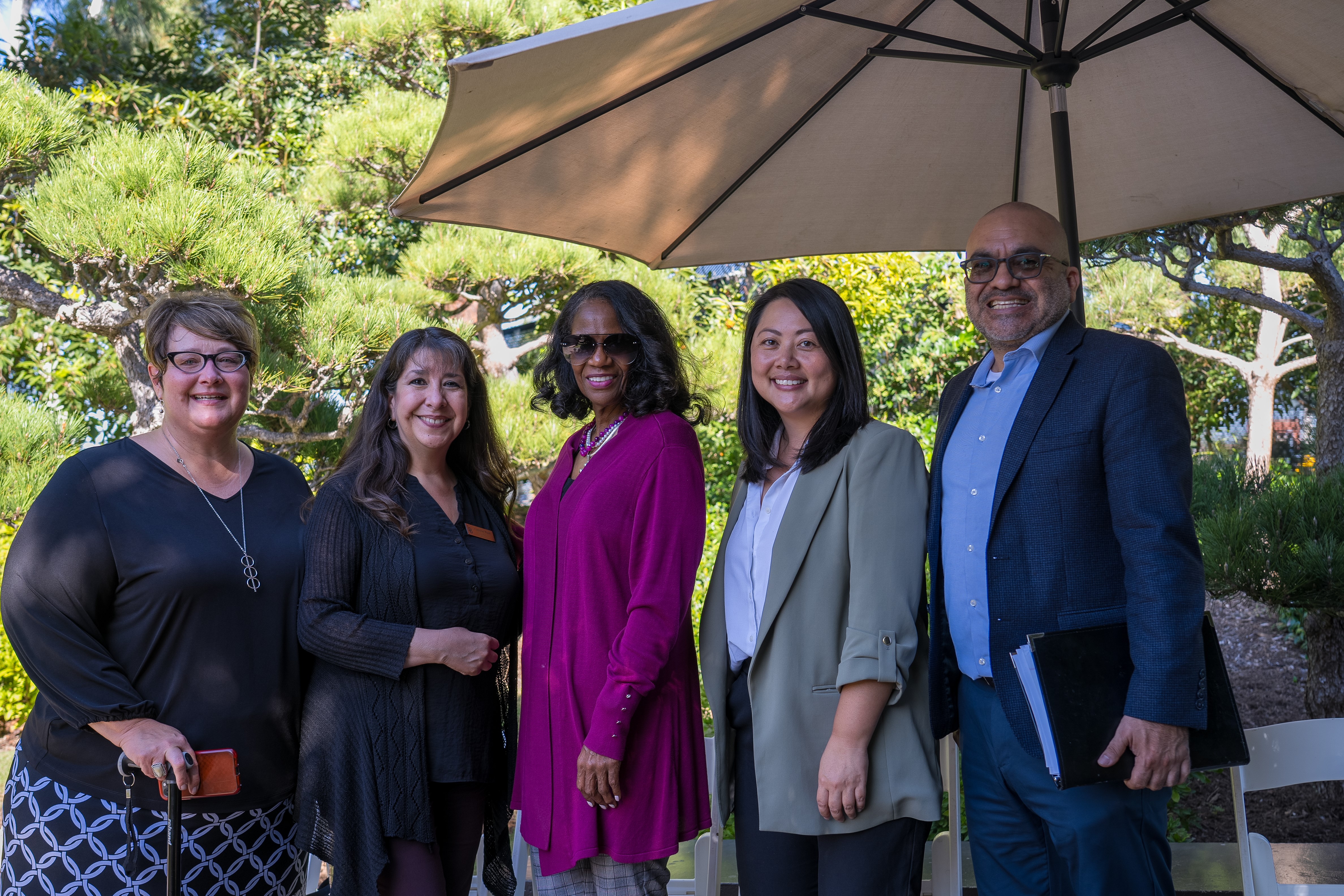 Five people stand under a patio umbrella in a garden