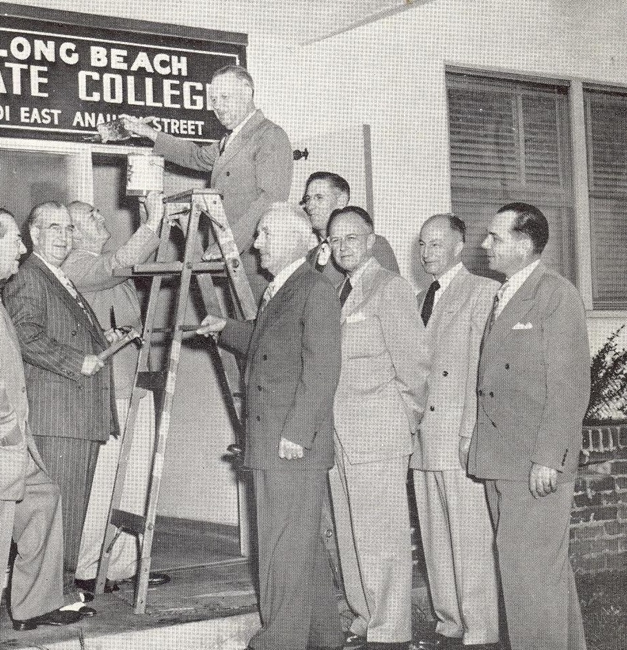 J. Victor Peterson hangs the "Long Beach State College" sign with other faculty A group of men in suits gathers around a man on a ladder, holding a paintbrush near a sign that reads "Long Beach State College."