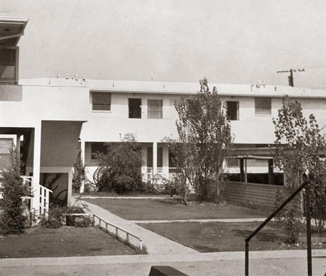 CSULB's first home, an apartment on Anaheim Road A two-story building with white walls and large windows stands in the background, with a small lawn and pathway leading toward it.