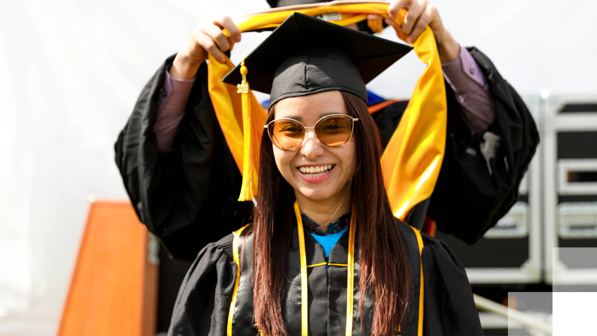 Beach EDGE program A graduate student in a black robe stands as someone behind her puts a graduate hood over her cap.