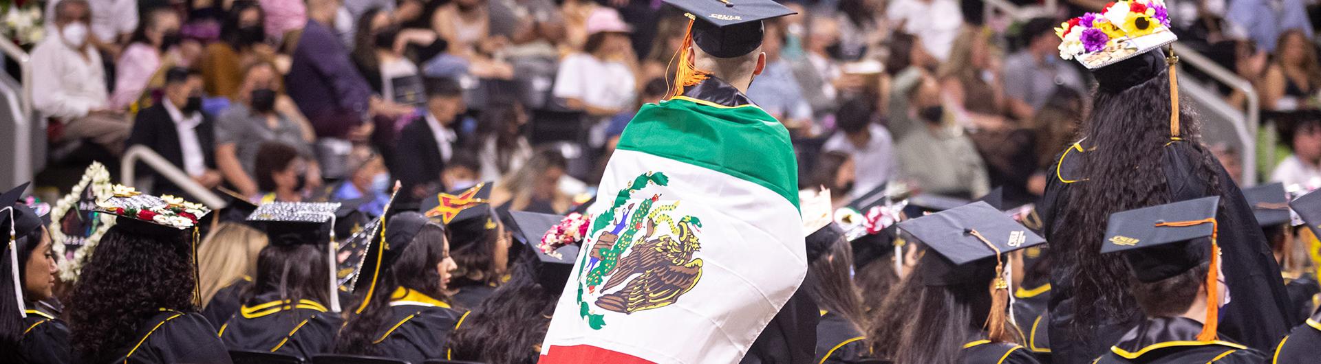 a Latine grad sitting in the stands at the cultural graduation ceremony holding the mexico flag