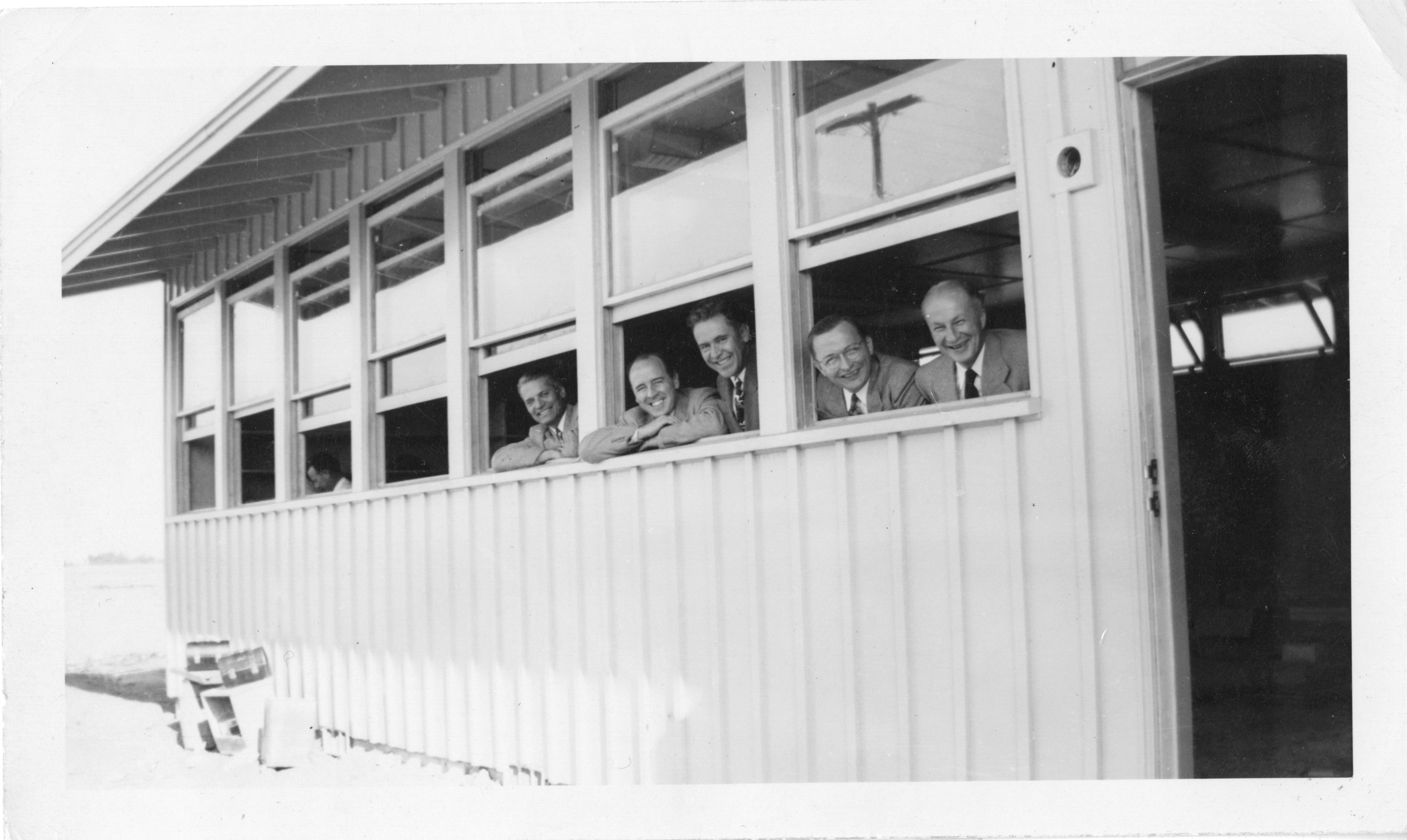 Faculty members look out of the original library building, 1949 Five men in suits lean out of windows, smiling from a building with horizontal paneling.
