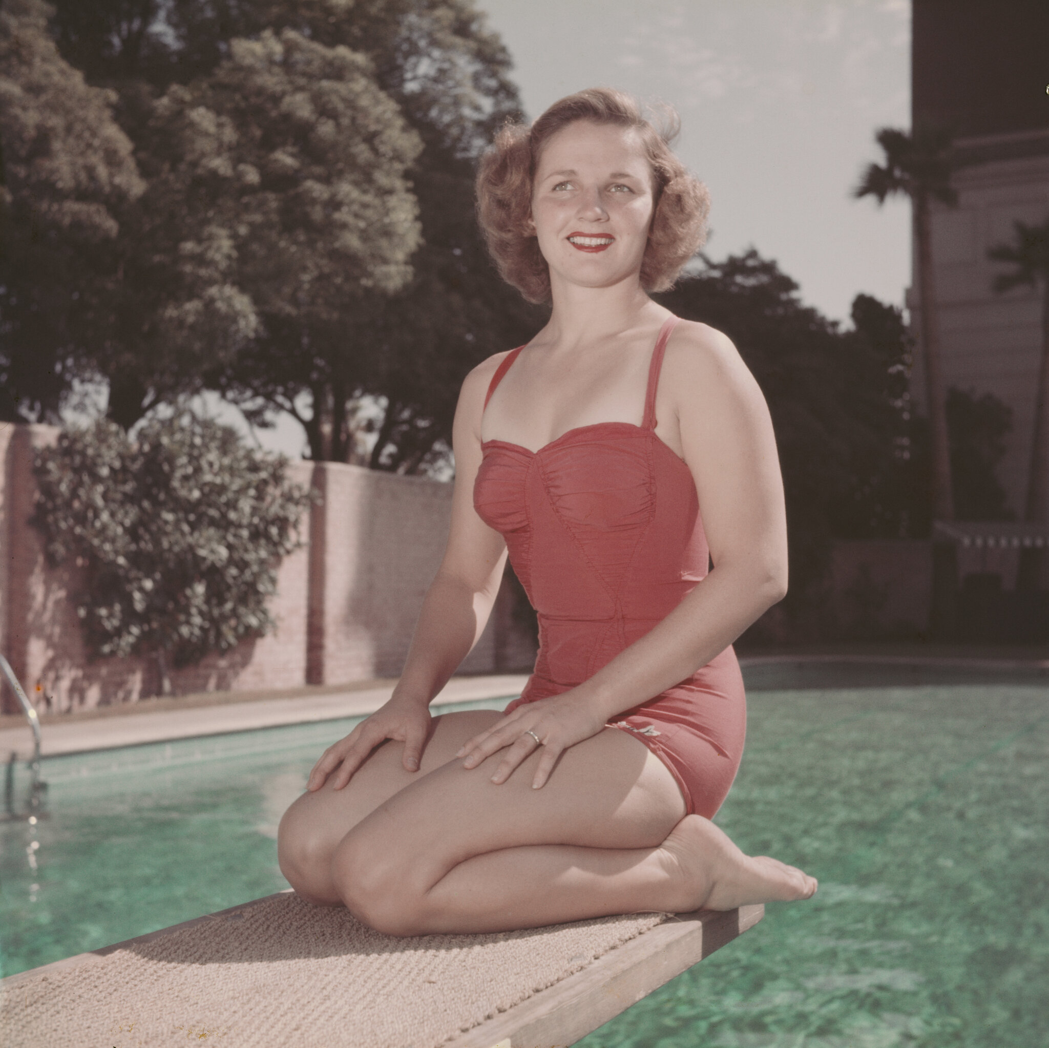 Pat McCormick A person in a red swimsuit kneels on a diving board by a pool, smiling at the camera.