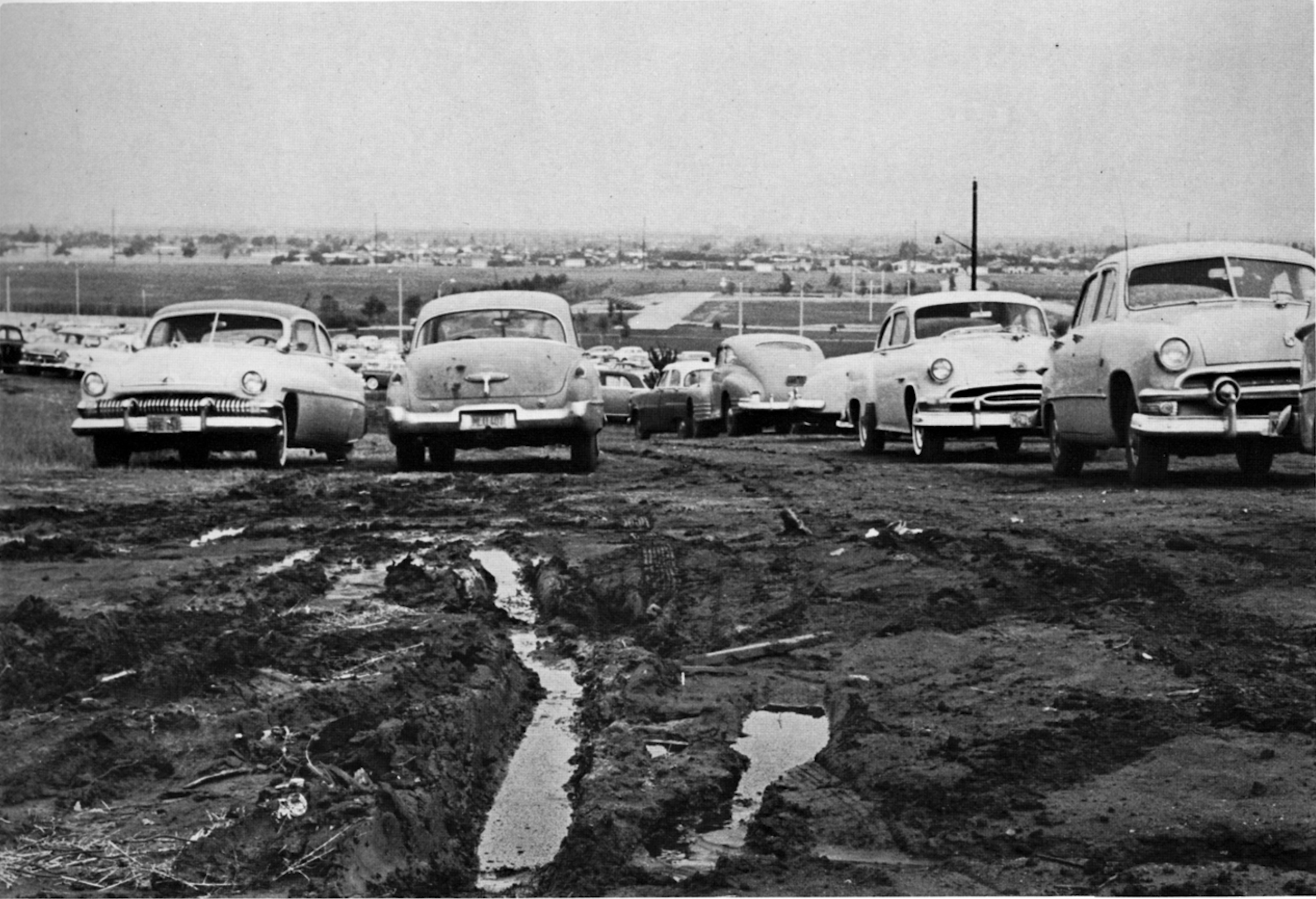 Cars are parked on a muddy dirt lot, early campus shot. Cars are parked on a muddy dirt lot, with deep tire tracks visible in the foreground.