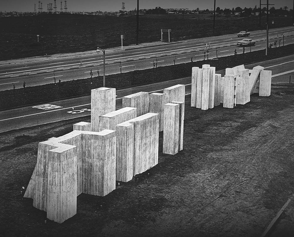 One of the sculptures built at CSULB A large concrete sculpture composed of block-like shapes stands near a road.