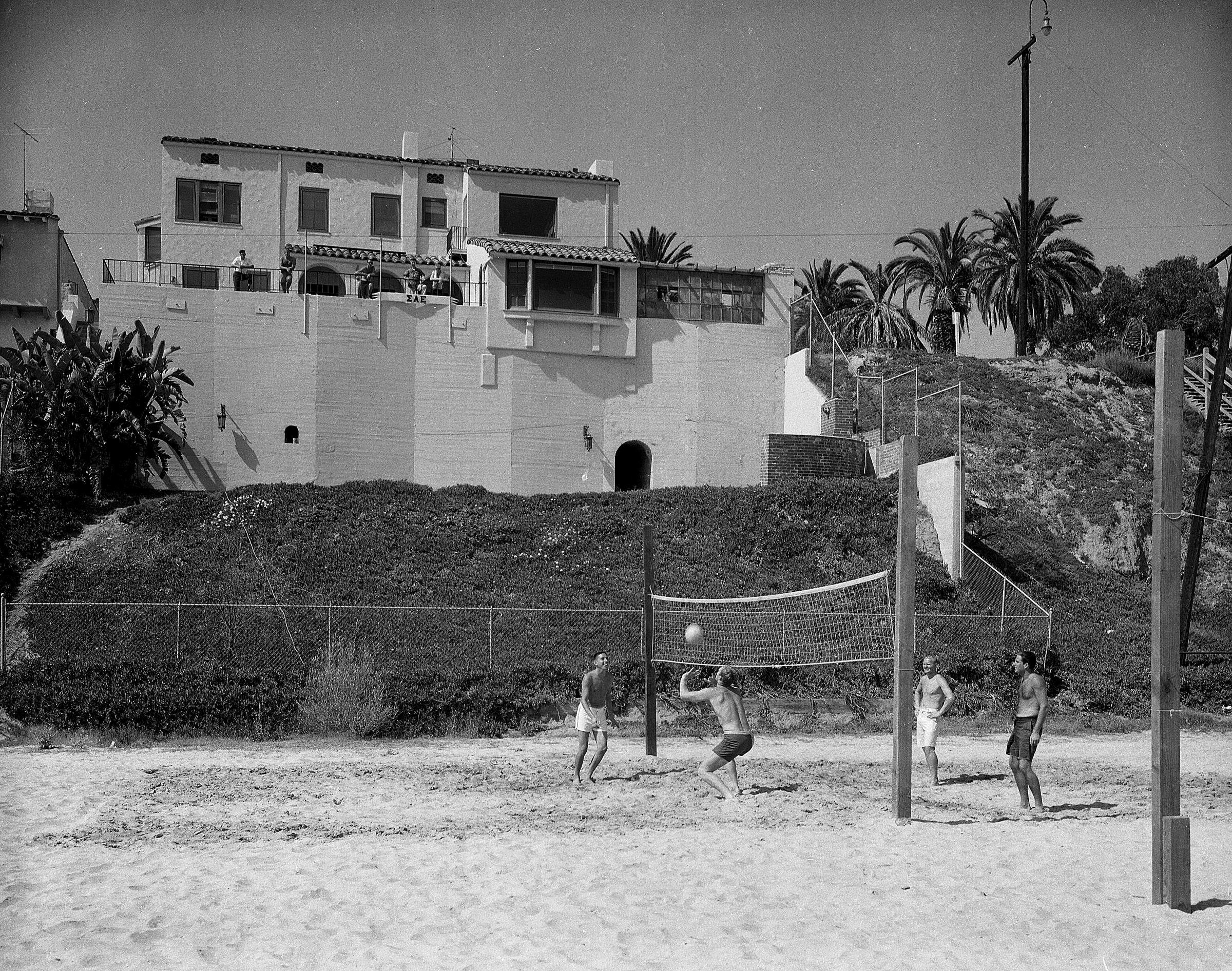 Sigma Alpha Epsilon’s oceanview house, circa 1970 Four people play beach volleyball near a large house overlooking the beach.
