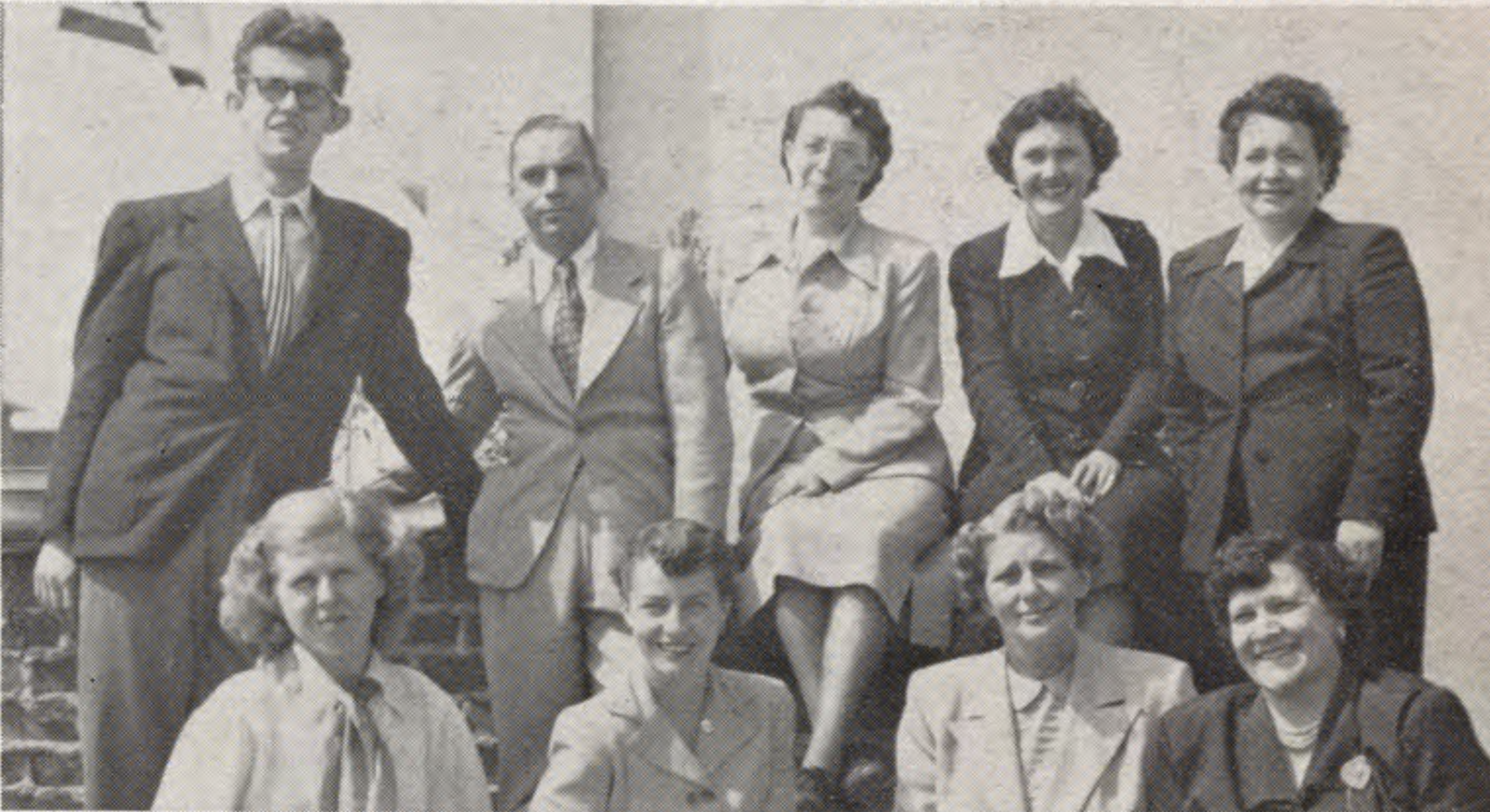 A black and white photo of nine students from the first class in 1950 A group of eight adults in formal attire poses for a photo outdoors, with five standing and three seated.