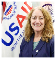 Karen Klimowski, DDIR Karen Klimowski, a white woman with blond, shoulder-length hair and a blue blazer, stands next to a USAID flag