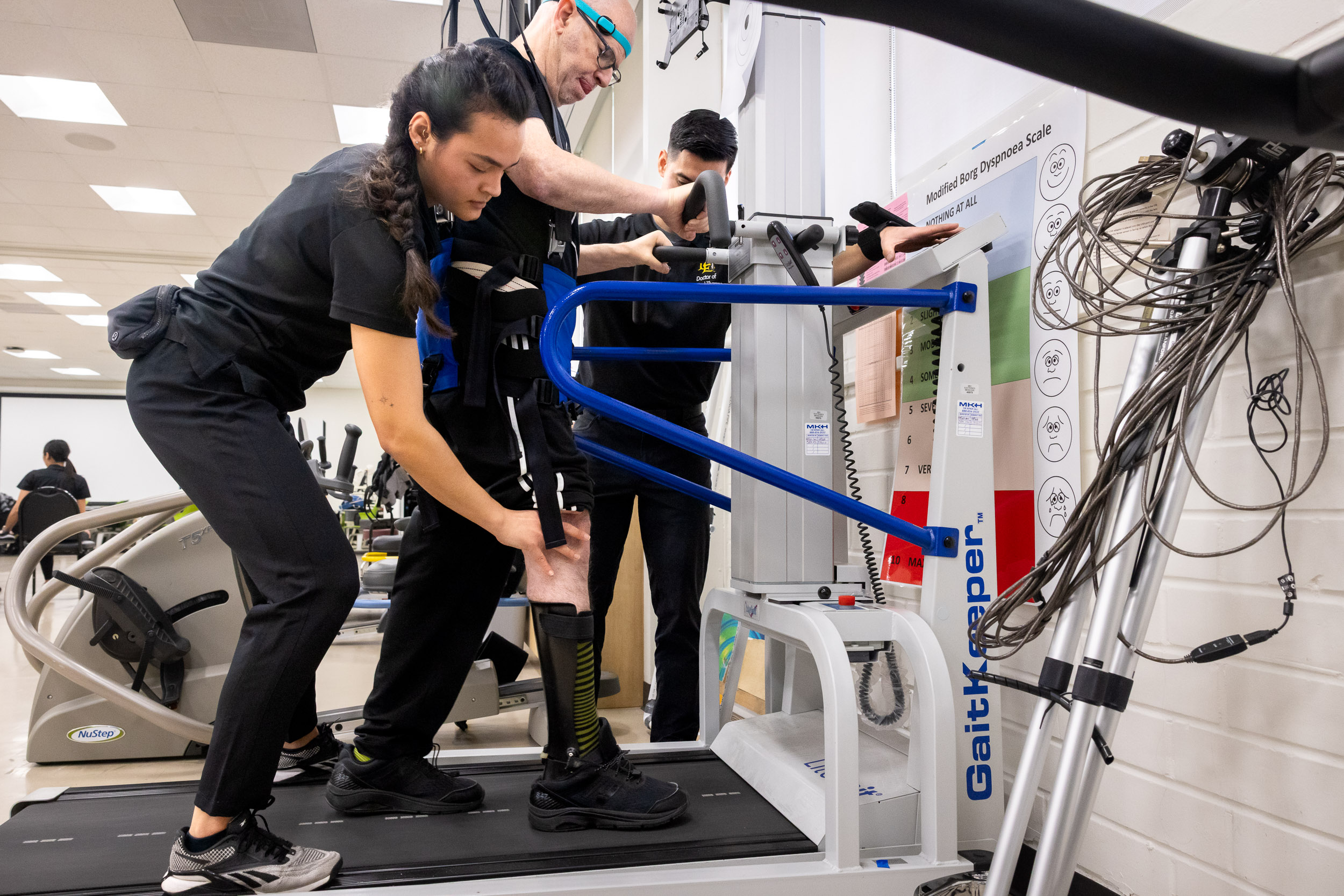 Romina Silva and another student help a Neuro Lab client with a prosthetic leg walk on a treadmill using a harness system