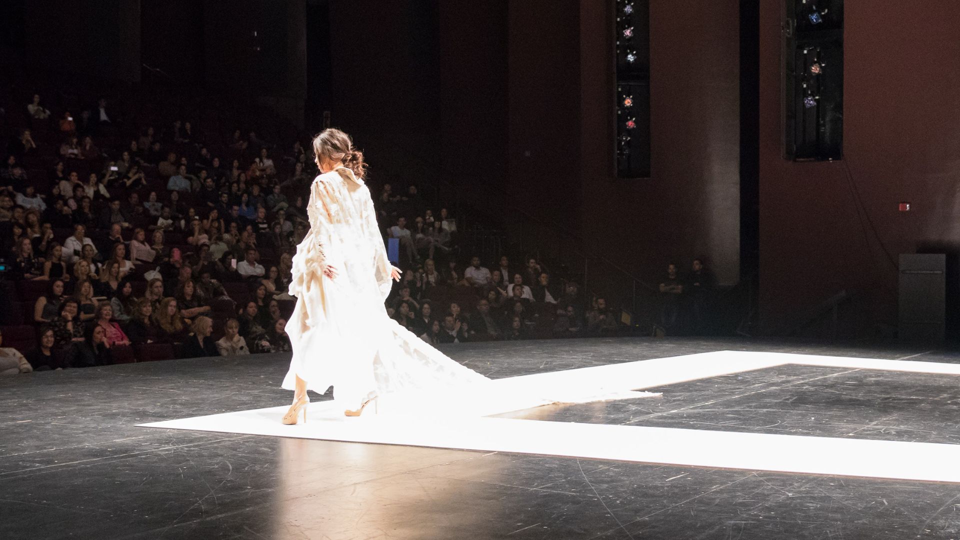 Model in a long white gown walks a lit runway before a seated audience. Model in a long white gown walks a lit runway before a seated audience.