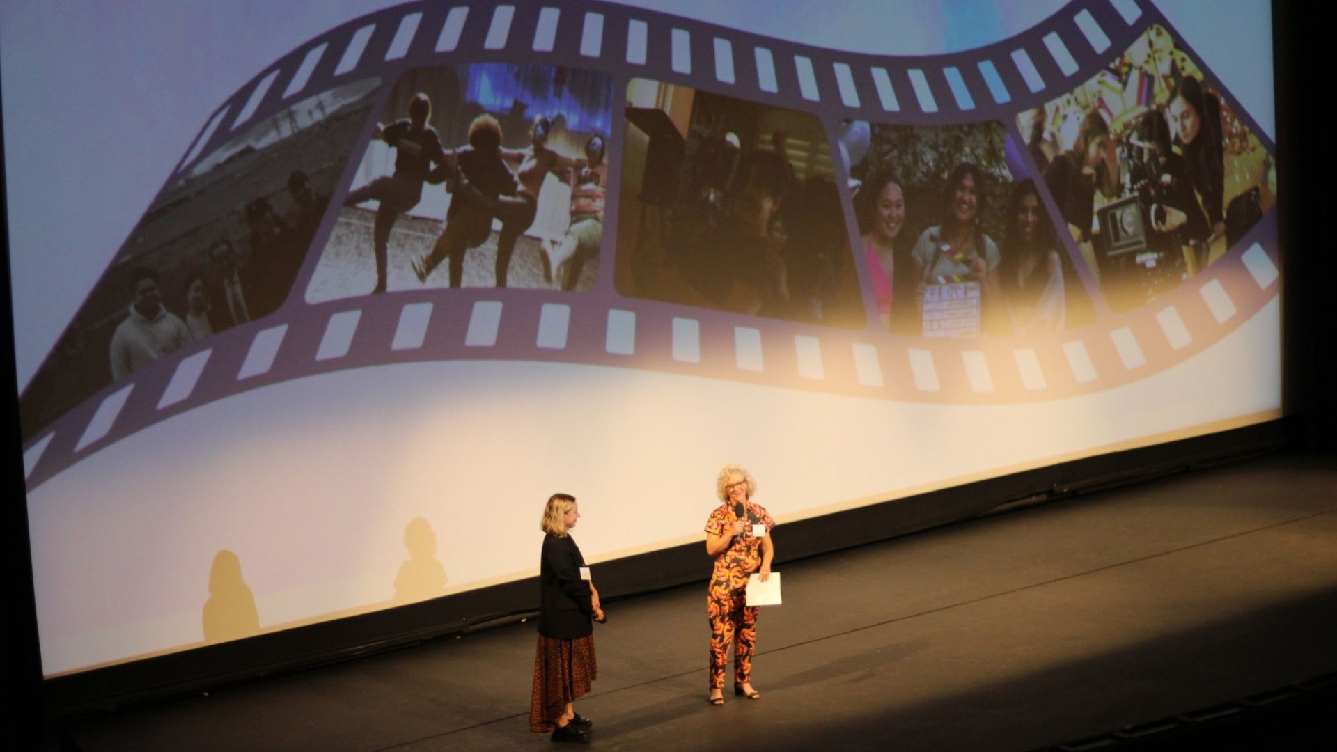Two people stand onstage speak at CSULB Film Festival Two people stand onstage speaking in front of a projected filmstrip display.