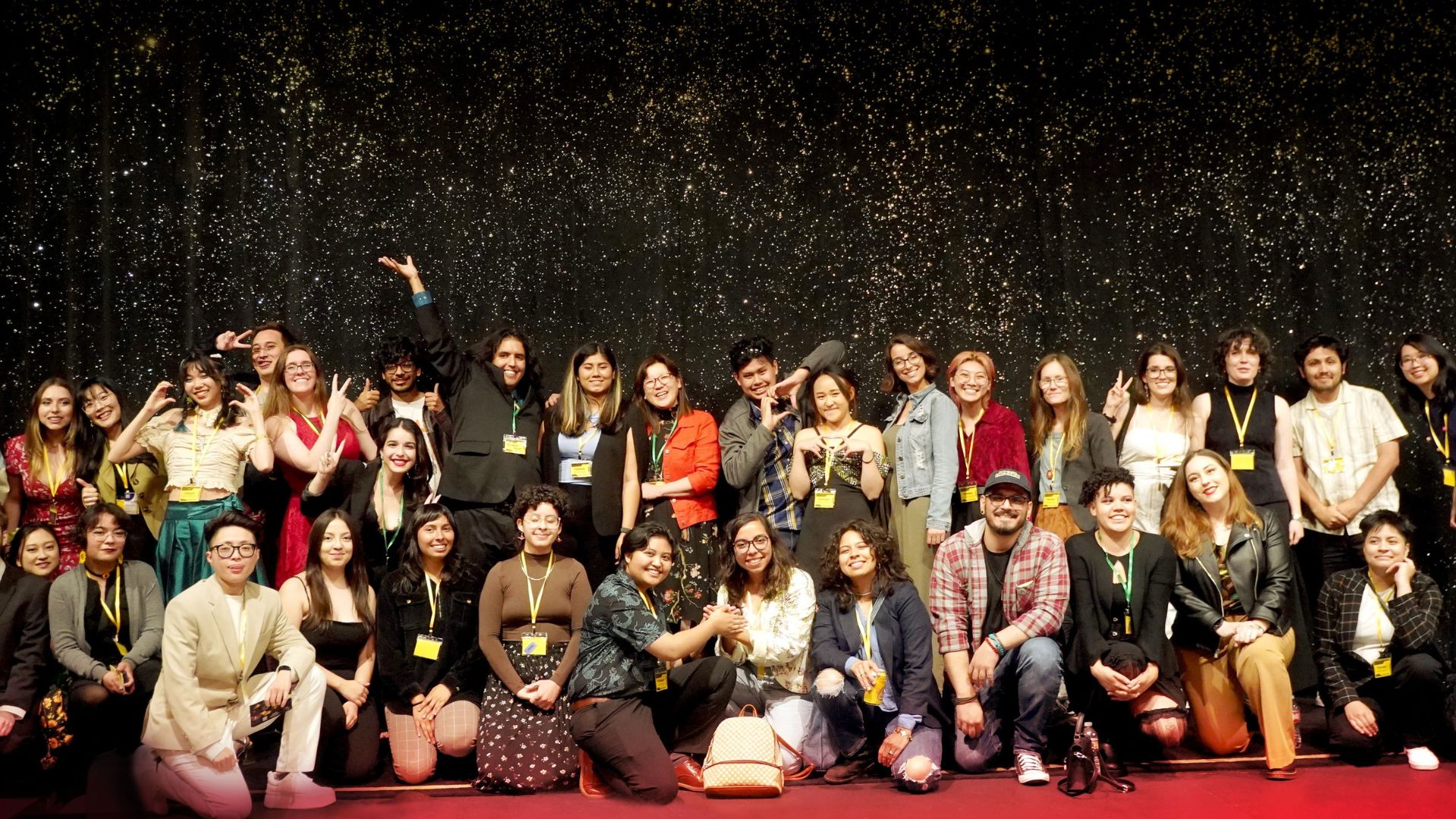 Large group poses in front of a glittery backdrop at CSULB Animation Festival Large group poses in front of a glittery backdrop wearing event badges and smiling.