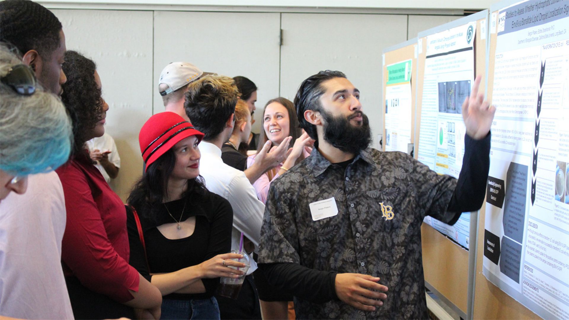 Presenter gestures toward a research poster as a crowd listens at RCSA Presenter gestures toward a research poster as a crowd listens and observes