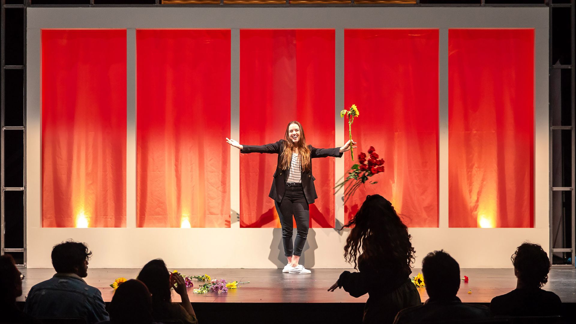 Performer at CSULB Theater holding flowers stands center stage smiling Performer holding flowers stands center stage smiling as audience members applaud.