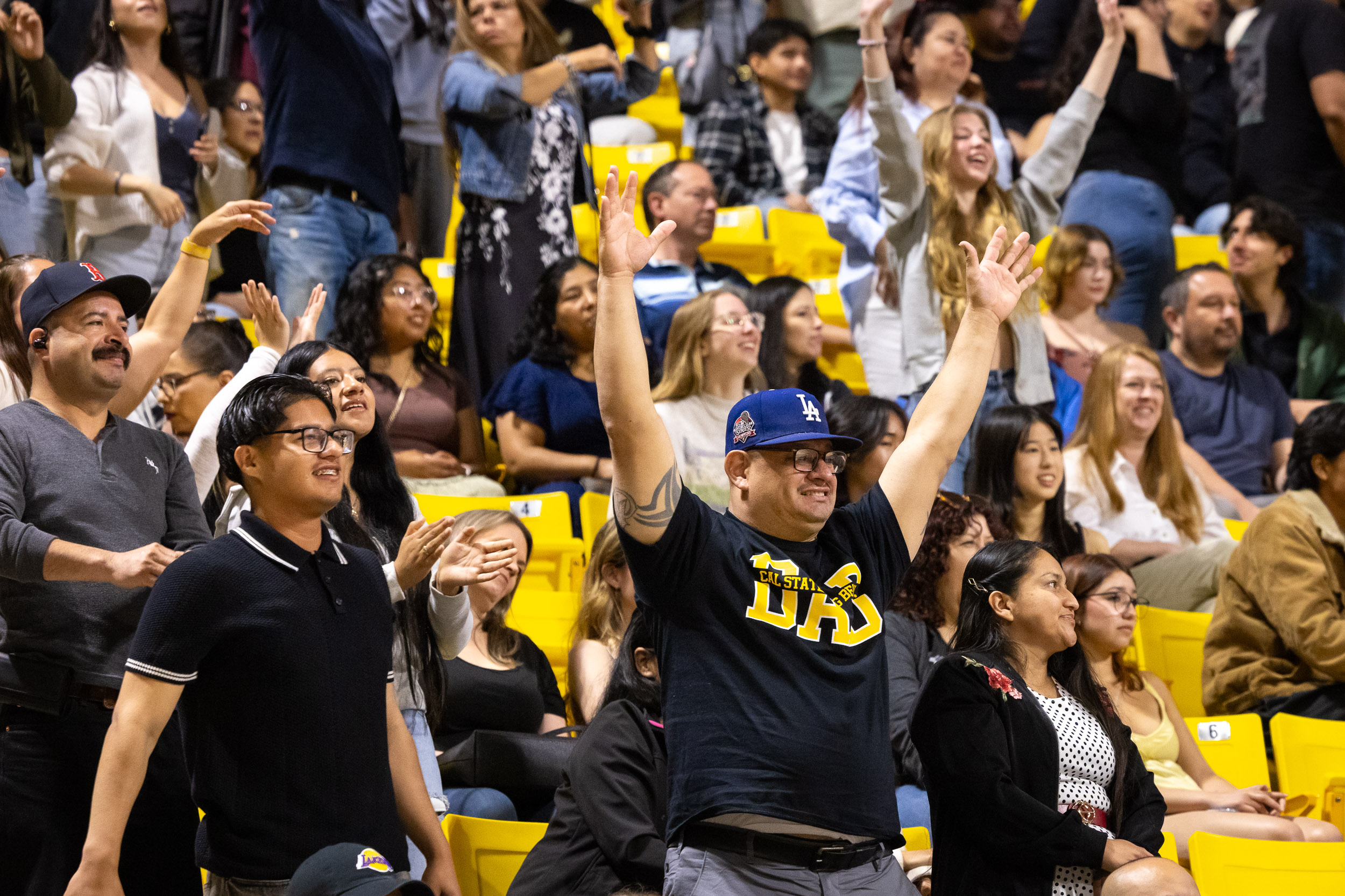 A cheering crowd fills yellow seats as a person in a Cal State Long Beach shirt raises both arms.