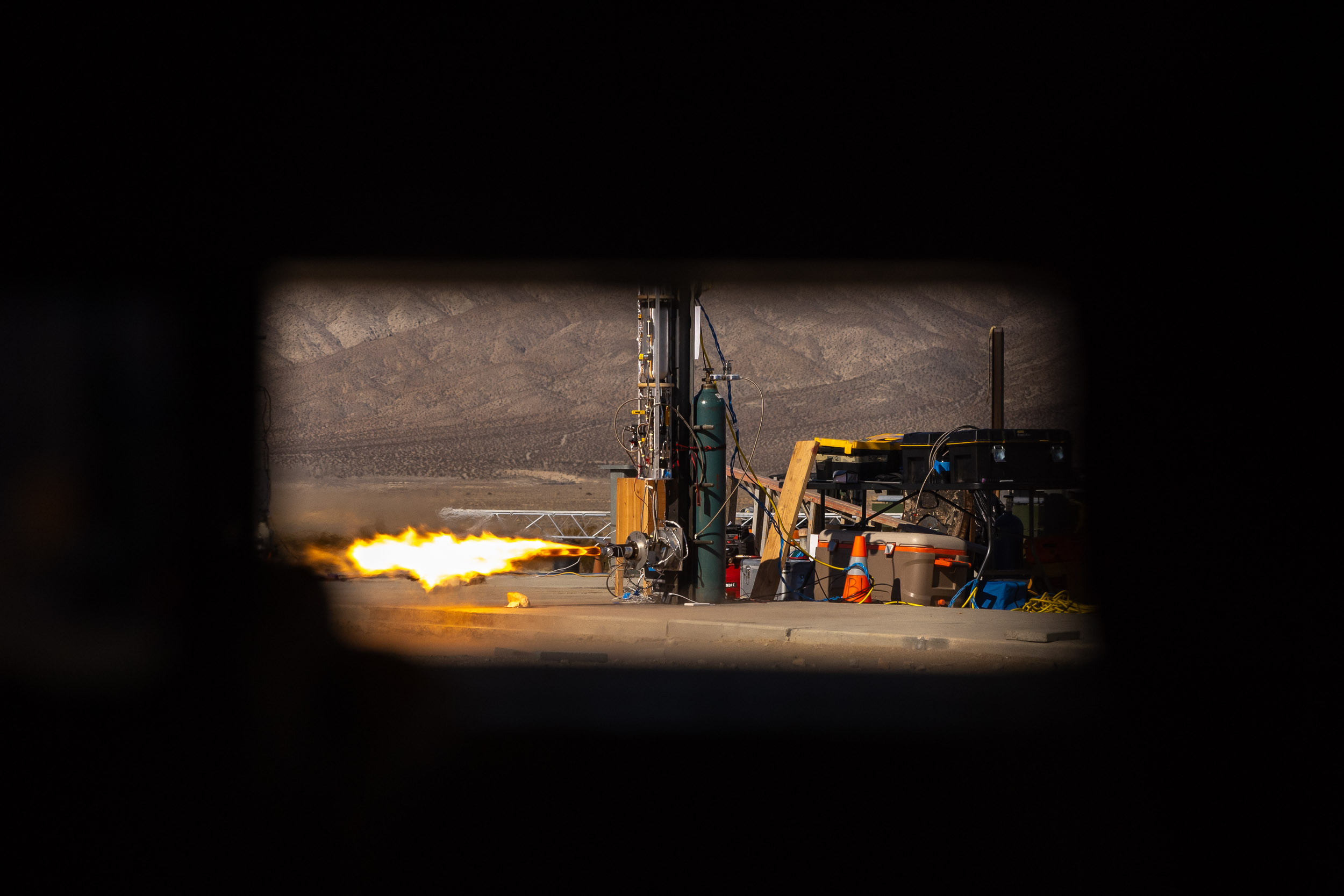 A rocket engine test fires on a stand, viewed through a narrow opening with flame visible