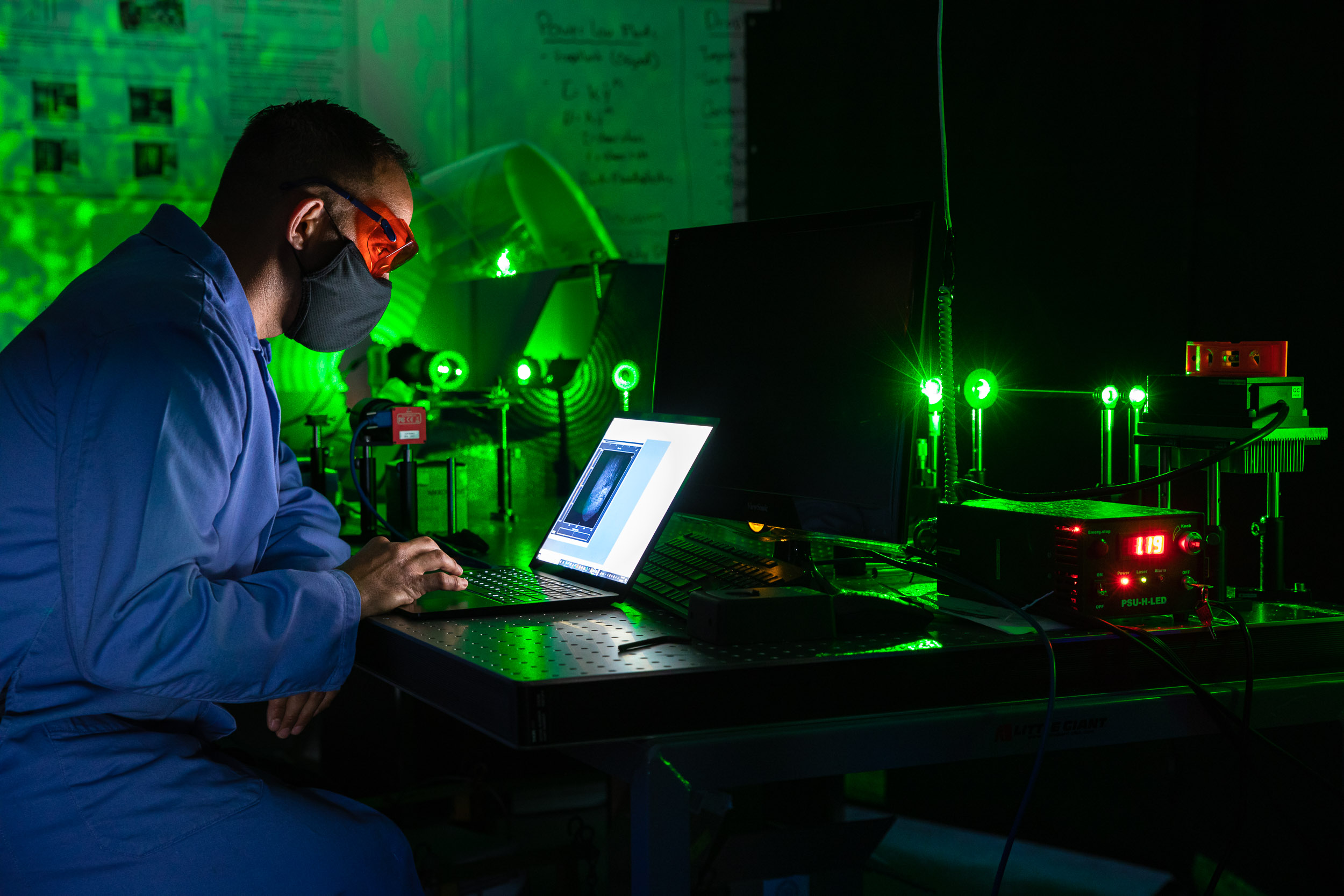 A person in safety gear analyzes data on a computer in a darkened optics lab