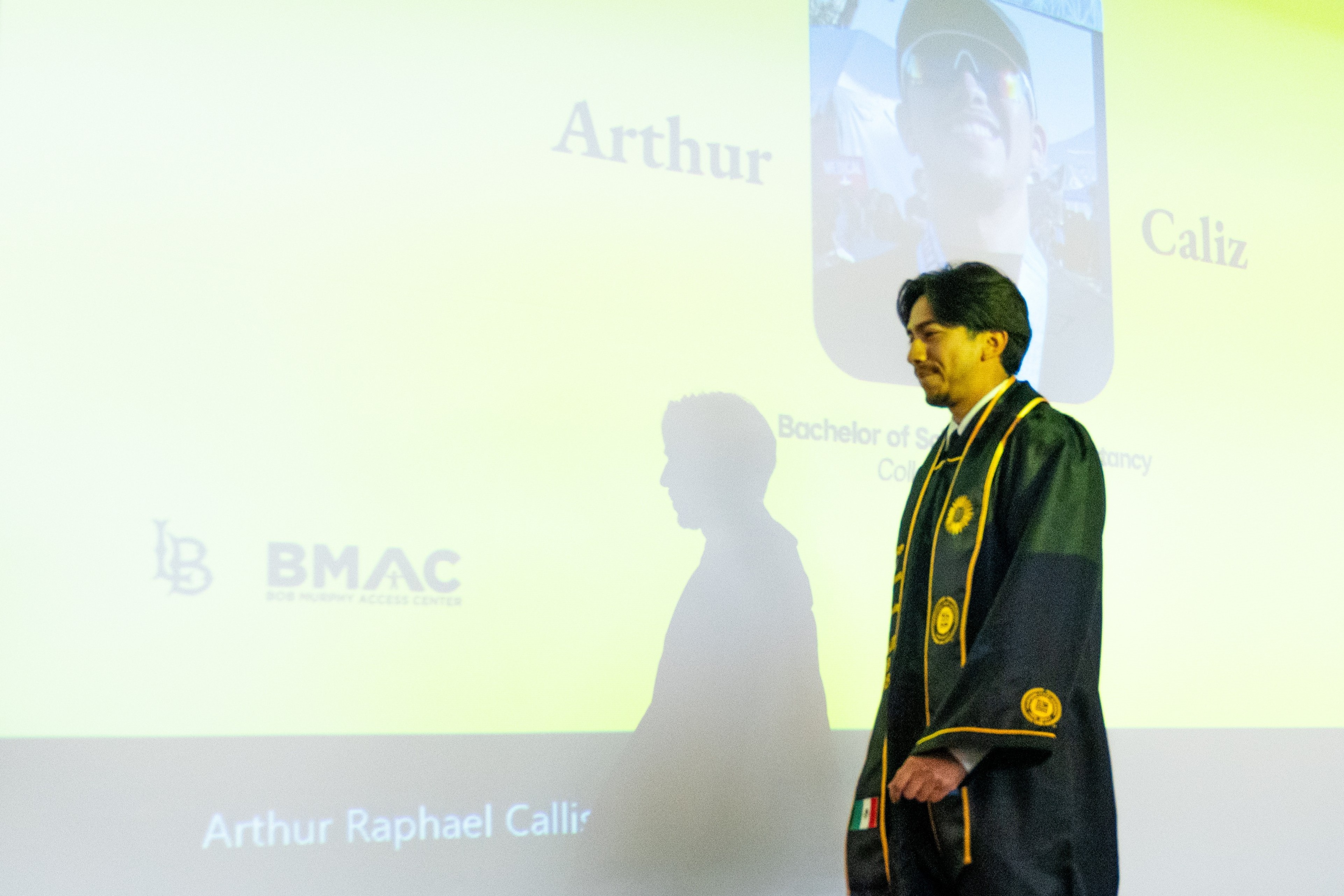 A male graduate in a black cap and gown with gold trim walks across a raised stage. Behind him, a large screen shows a yellow slide with his name “Arthur Caliz” and his photo.