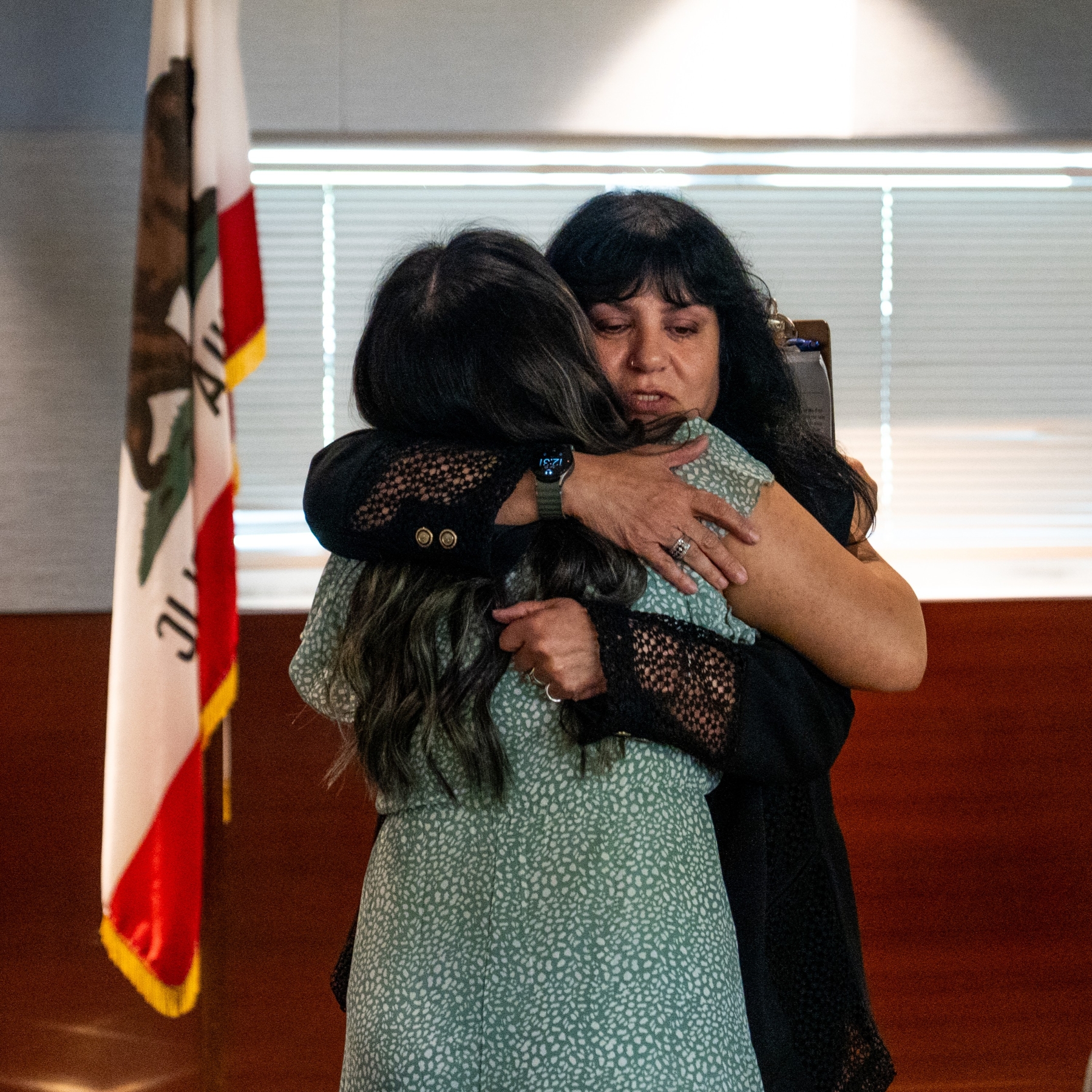 Two women embrace indoors. The woman facing the camera wears black clothing with a watch and rings; the other wears a light green, patterned dress. A California state flag stands beside them and horizontal blinds cast soft light over the scene.