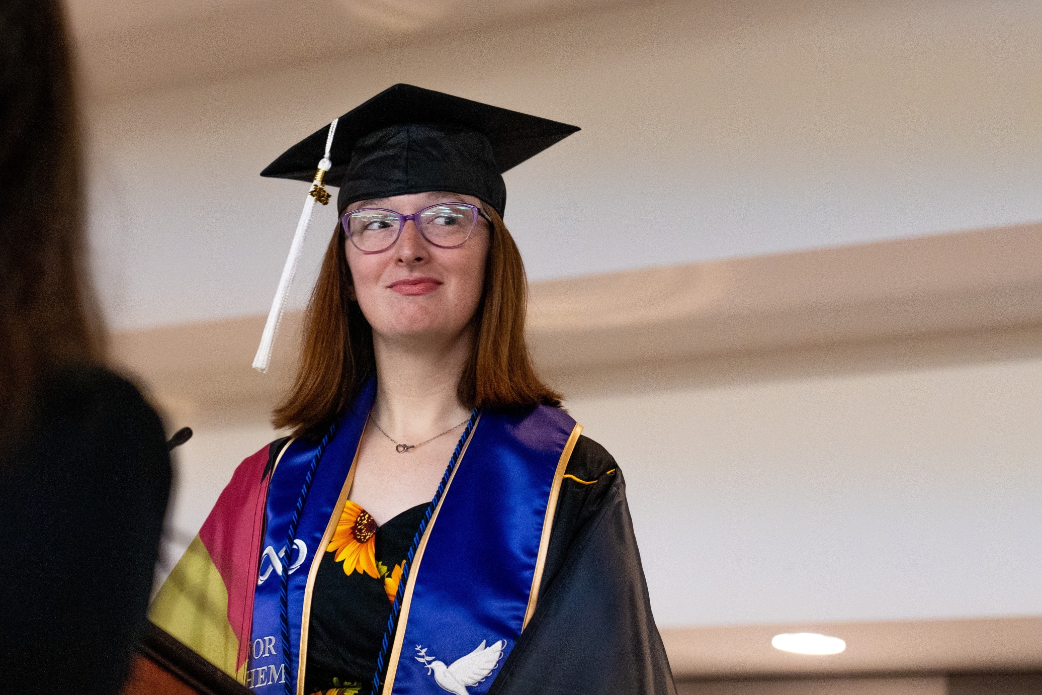 Clad in a black graduation cap and gown, the graduate stands looking to the right, wearing purple-framed glasses, a delicate necklace, and a blue-and-gold sash over a floral dress, with a white tassel hanging from the cap.