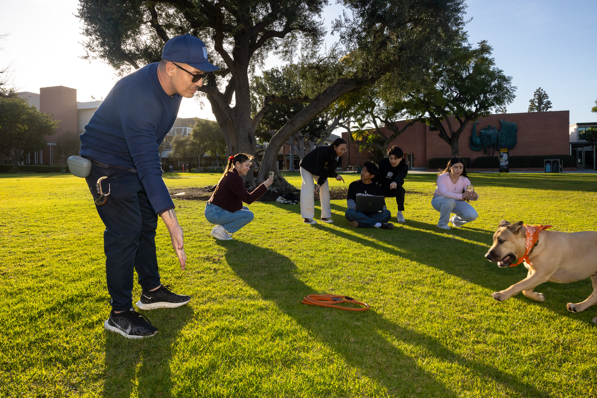A man plays with a dog in a grassy area while students take pictures on cell phones