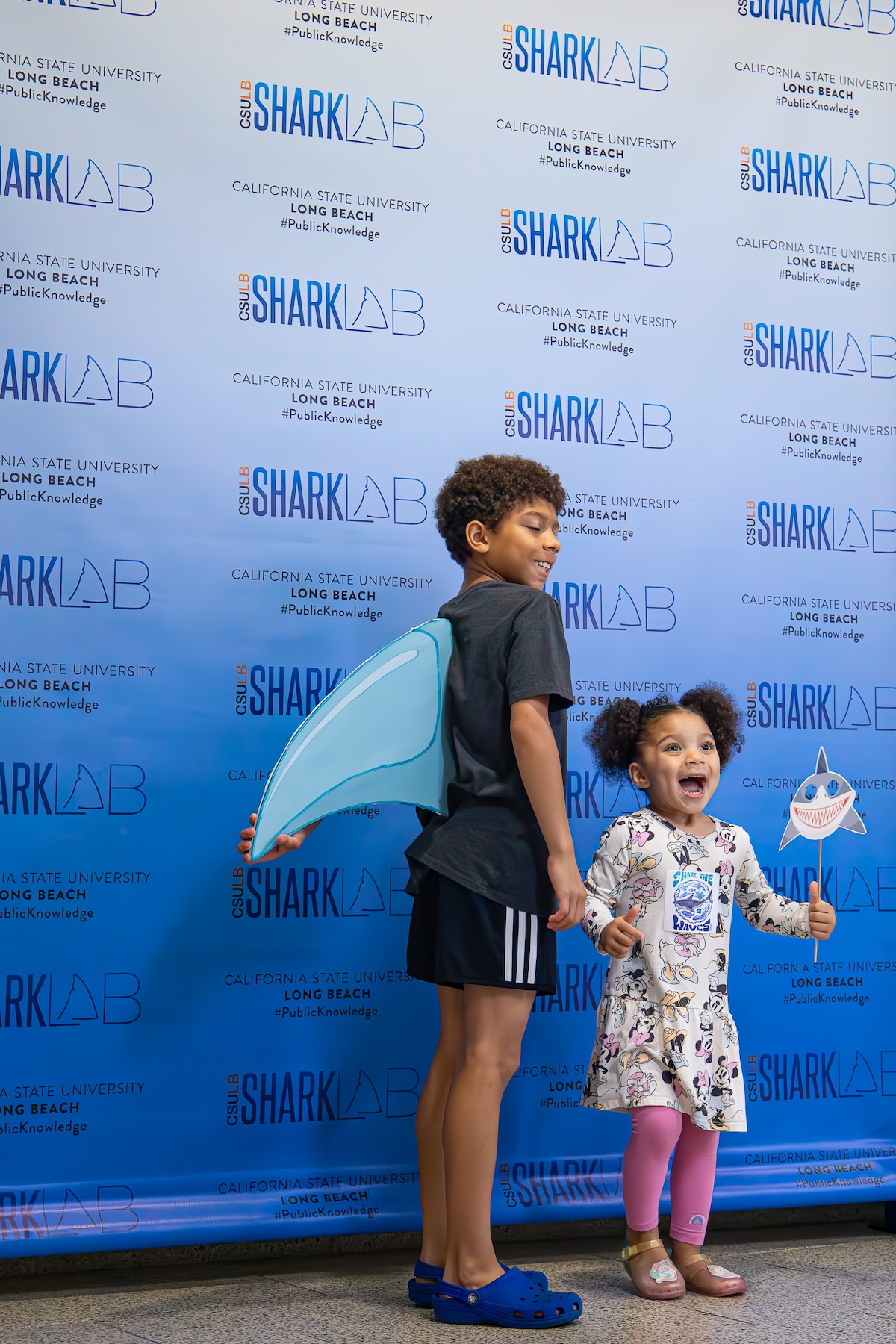 Two children pose in front of a CSULB Shark Lab backdrop with props
