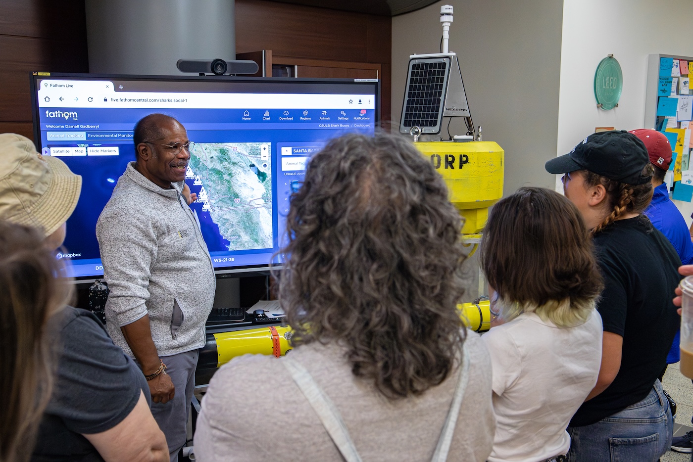 A person explains ocean tracking using a map on a screen to a group indoors