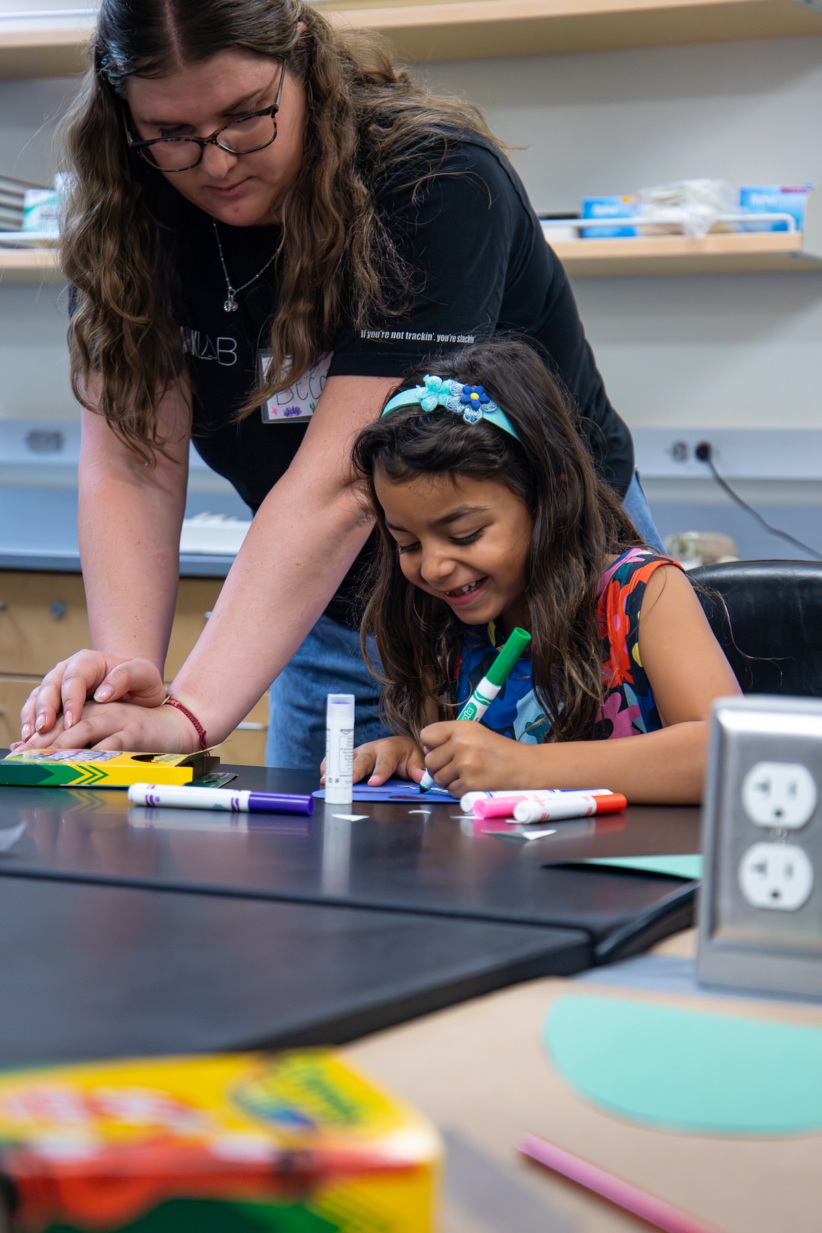 A child draws with markers at a table while a person helps guide their work