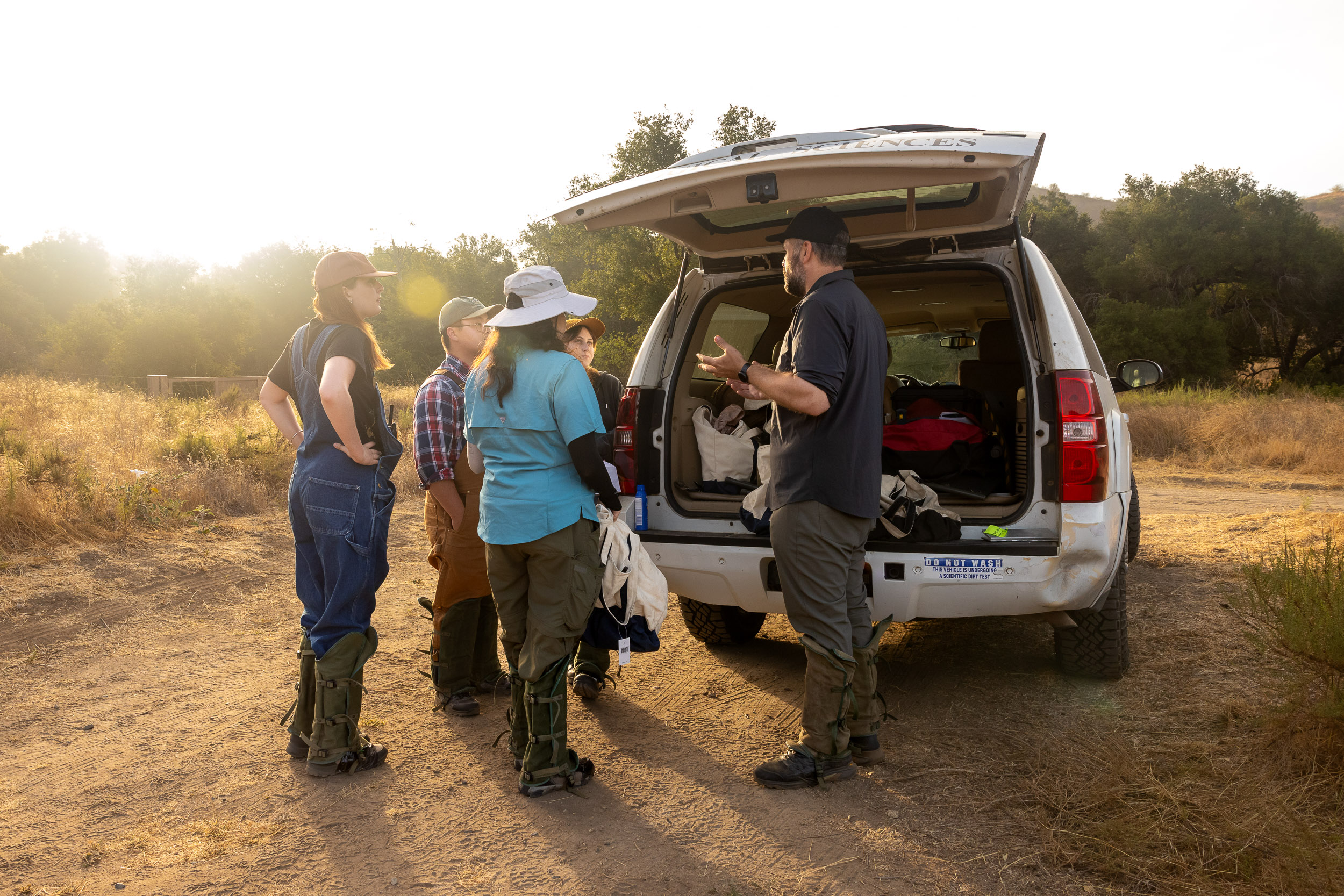 A man talks with students beside an open SUV in a sunlit field.