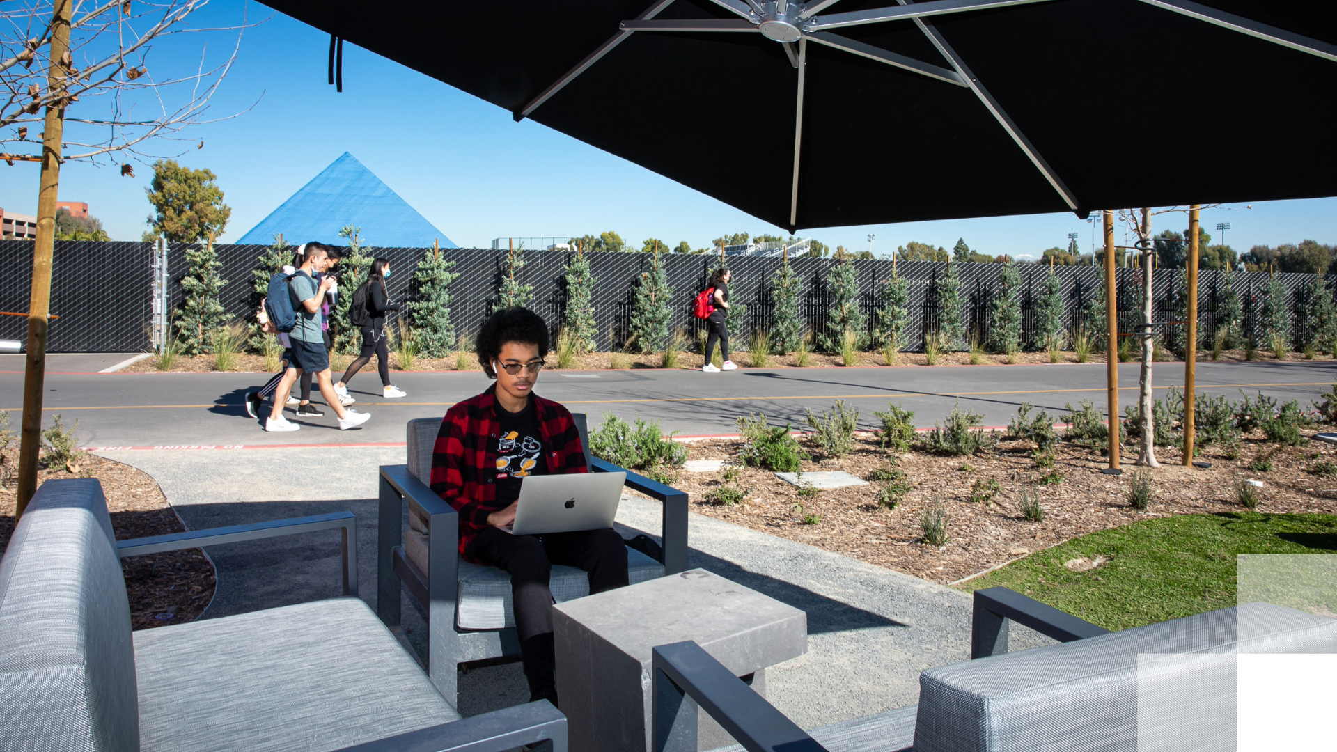 Build and Equitable and Empowering Culture Student working on a laptop under a patio umbrella with others walking nearby