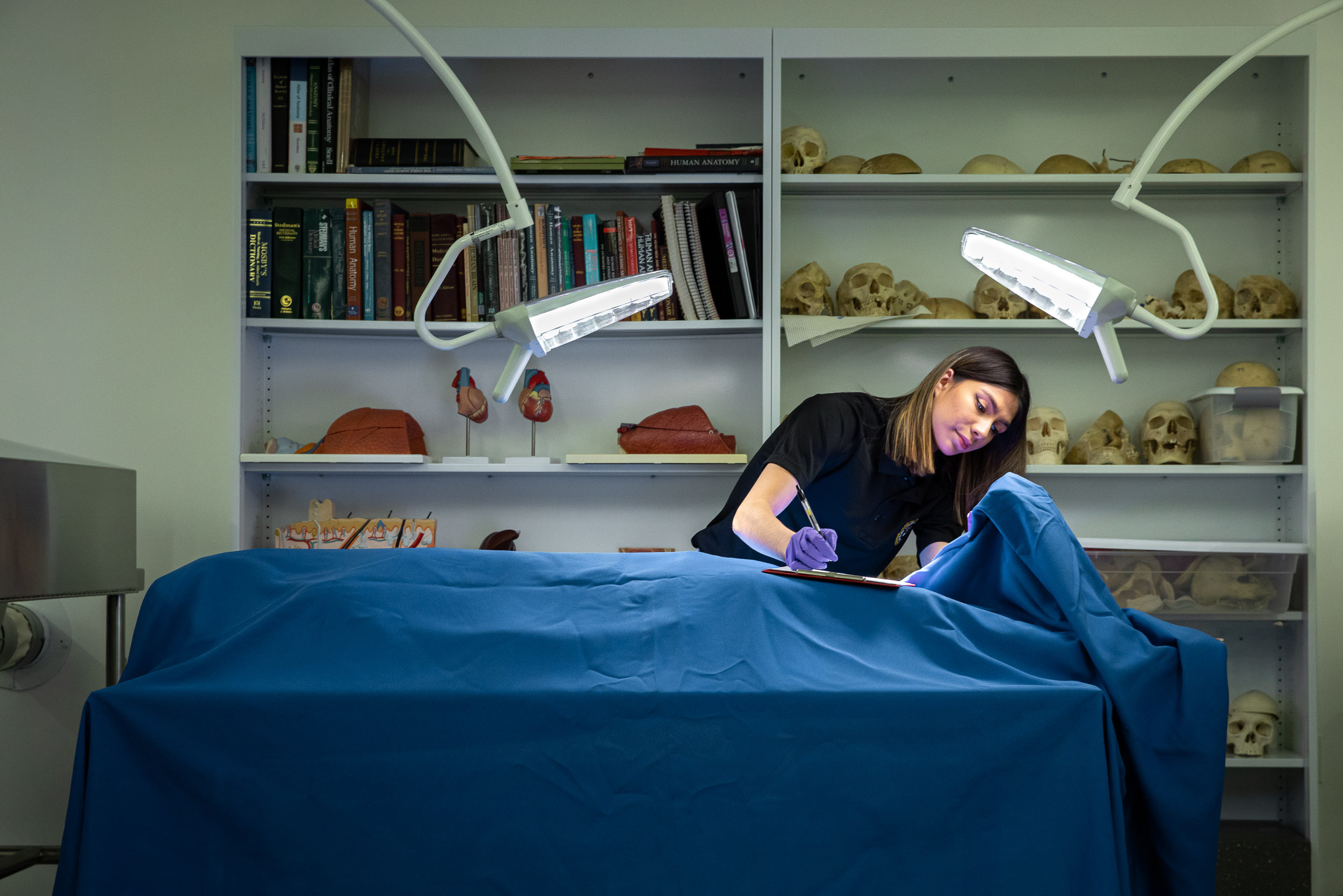 A person in gloves writes on a pad while inspecting a body under a sheet