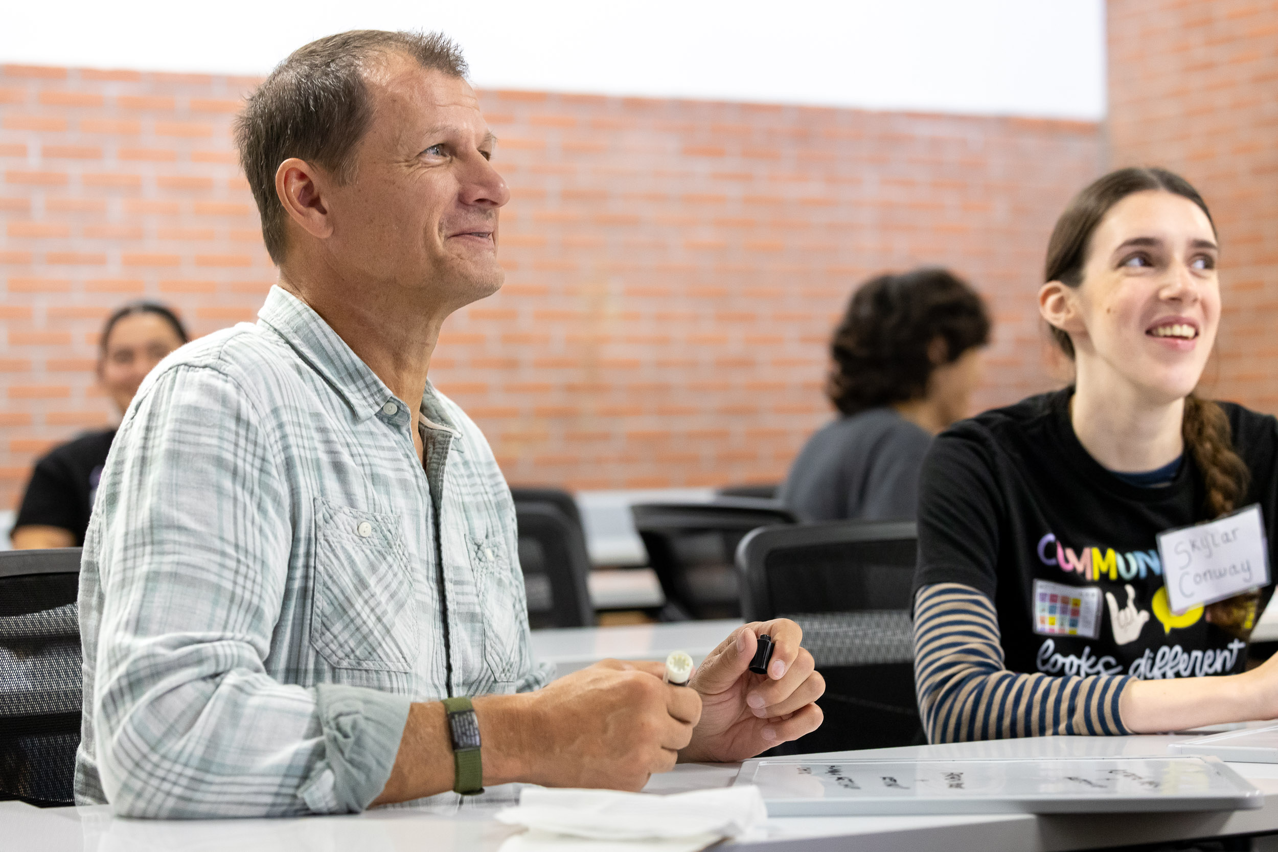 Two people sit at a table with whiteboards and listen during a discussion