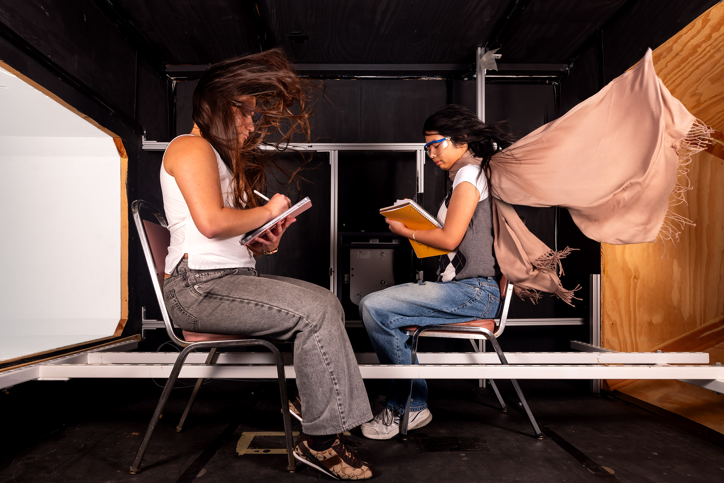 Two students sit facing each other in a wind tunnel with hair and clothing blowing as they write in notebooks