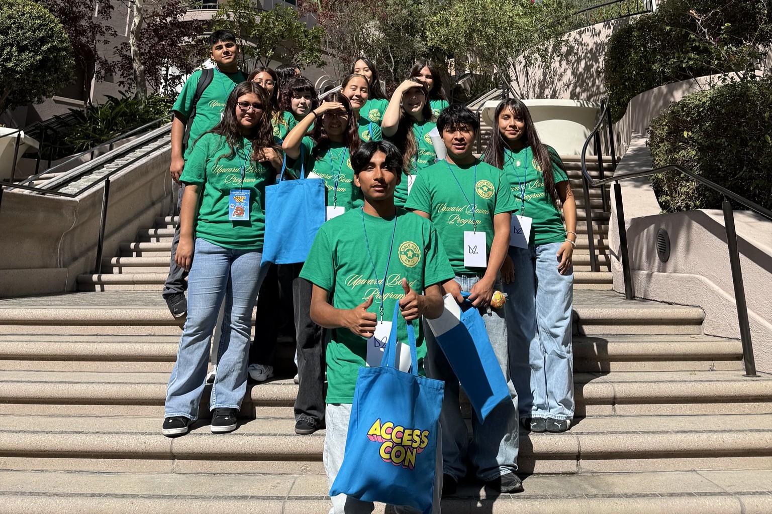 A group of students wearing matching green shirts pose on outdoor steps, smiling and holding blue tote bags.