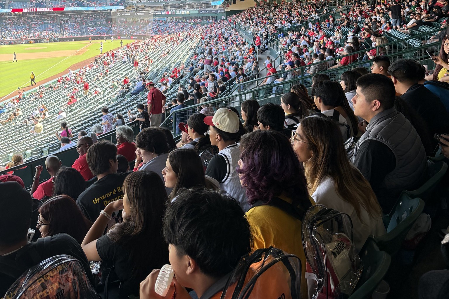 A group of students sits together in stadium seats watching a baseball game, with many spectators in the background.