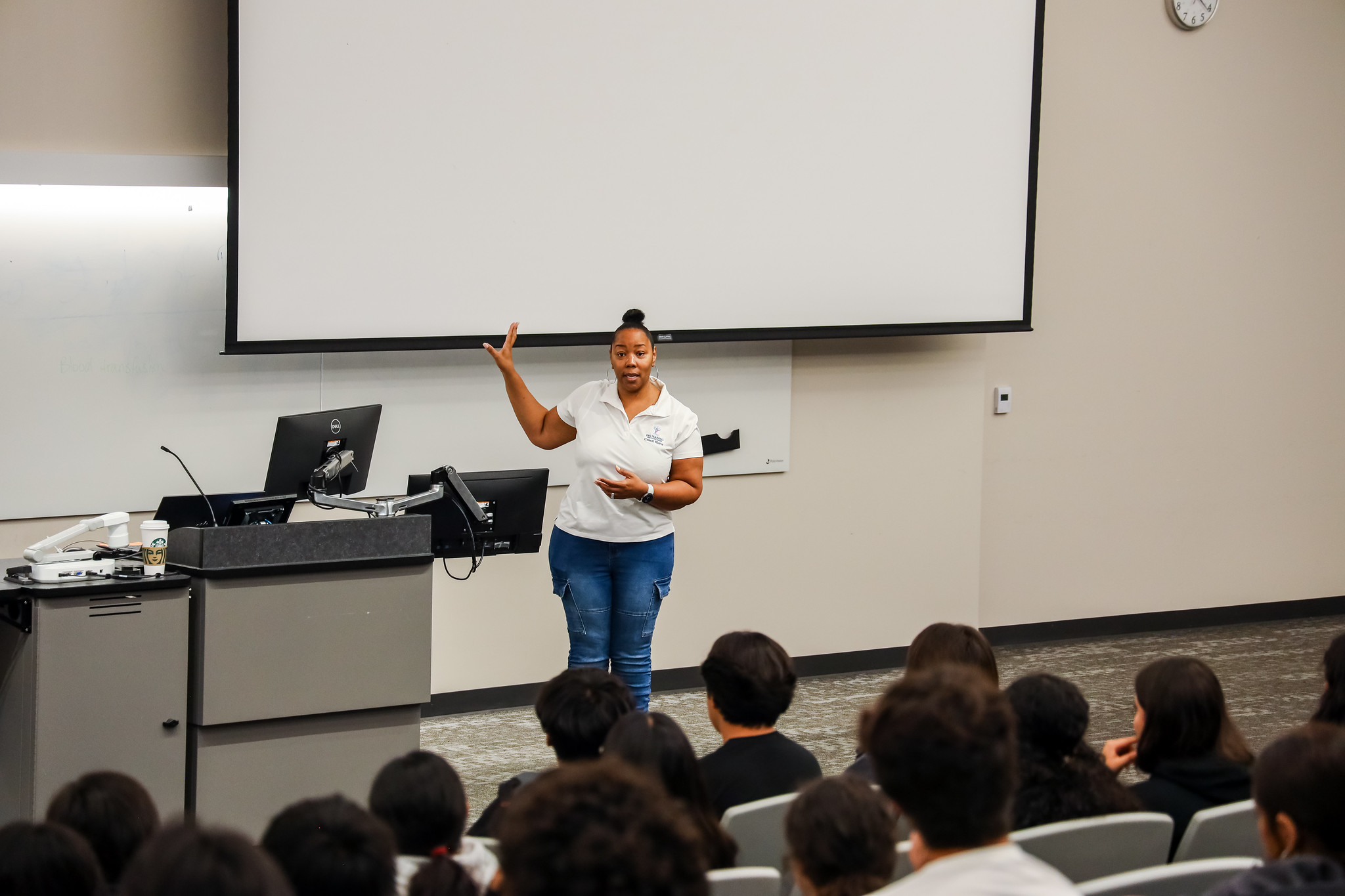 A presenter stands at the front of a classroom, gesturing while speaking to a group of seated students.