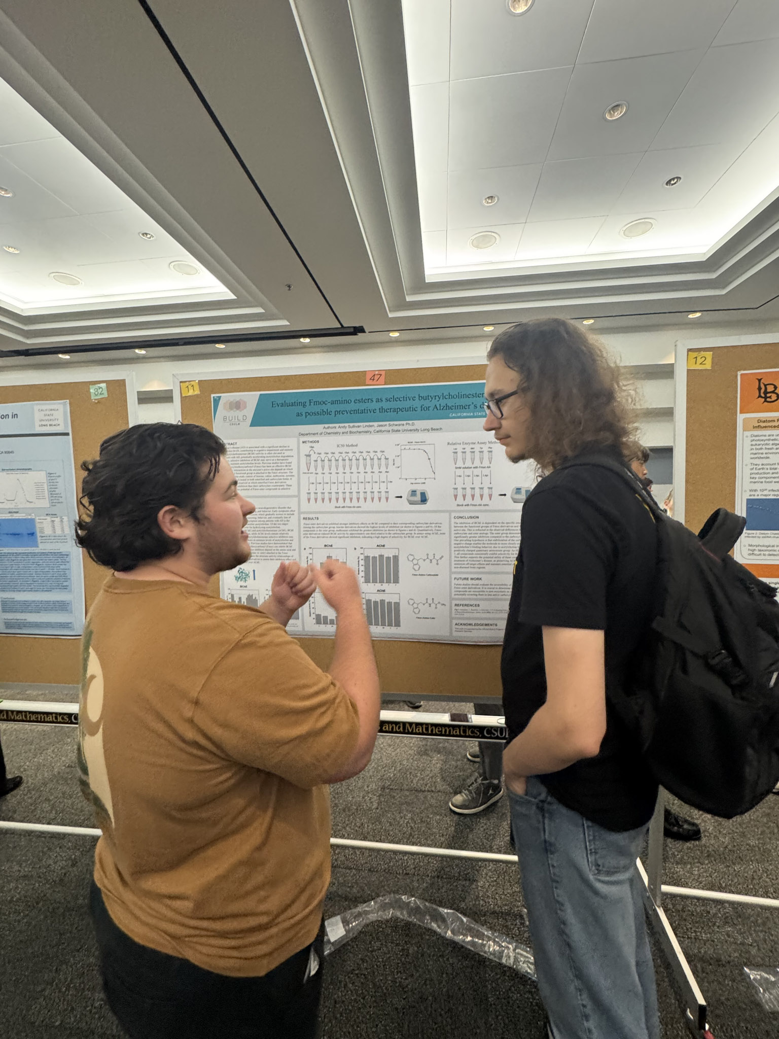 Presenter with black hair and brown shirt speaking with guest in front of poster