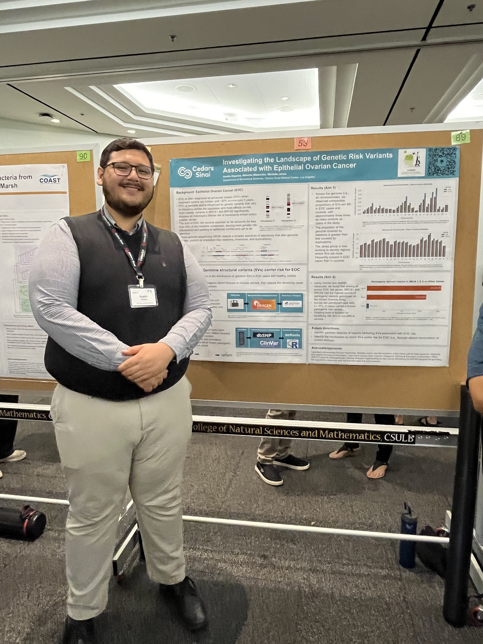 Male presenter with short brown hair and sweater vest over shirt standing in front of research poster