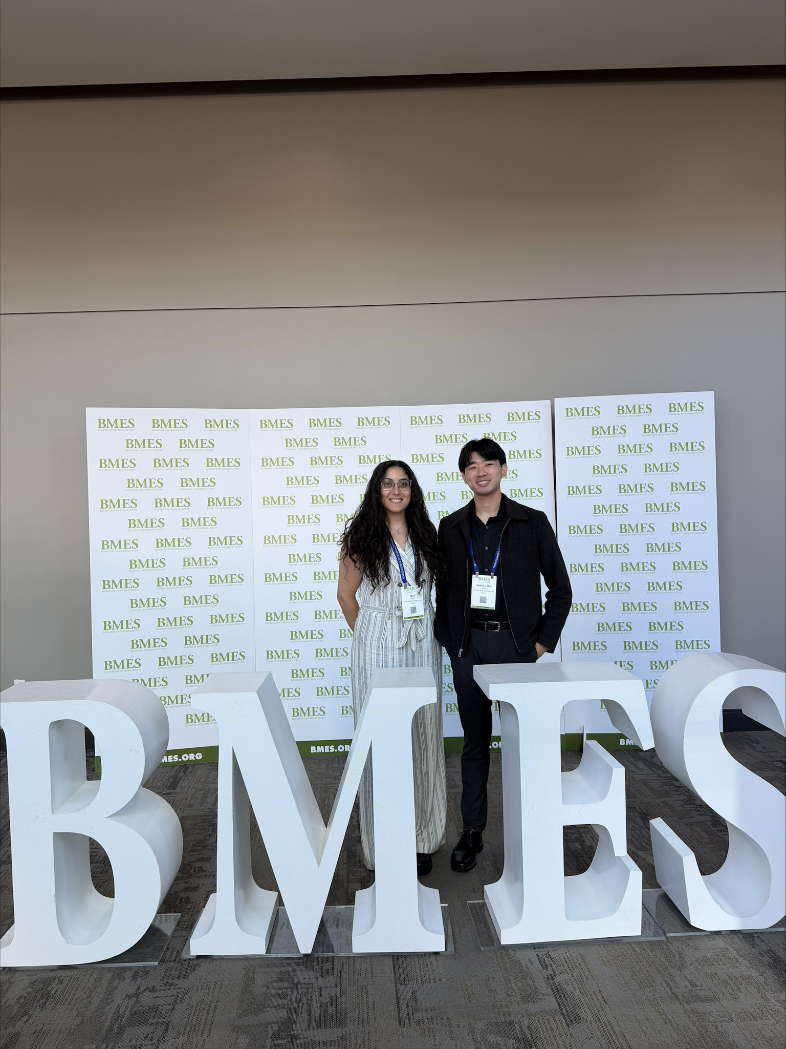 Woman with dark long hair and light outfit standing next to man with dark short hair and dark outfit; both behind letters that say "BMES"