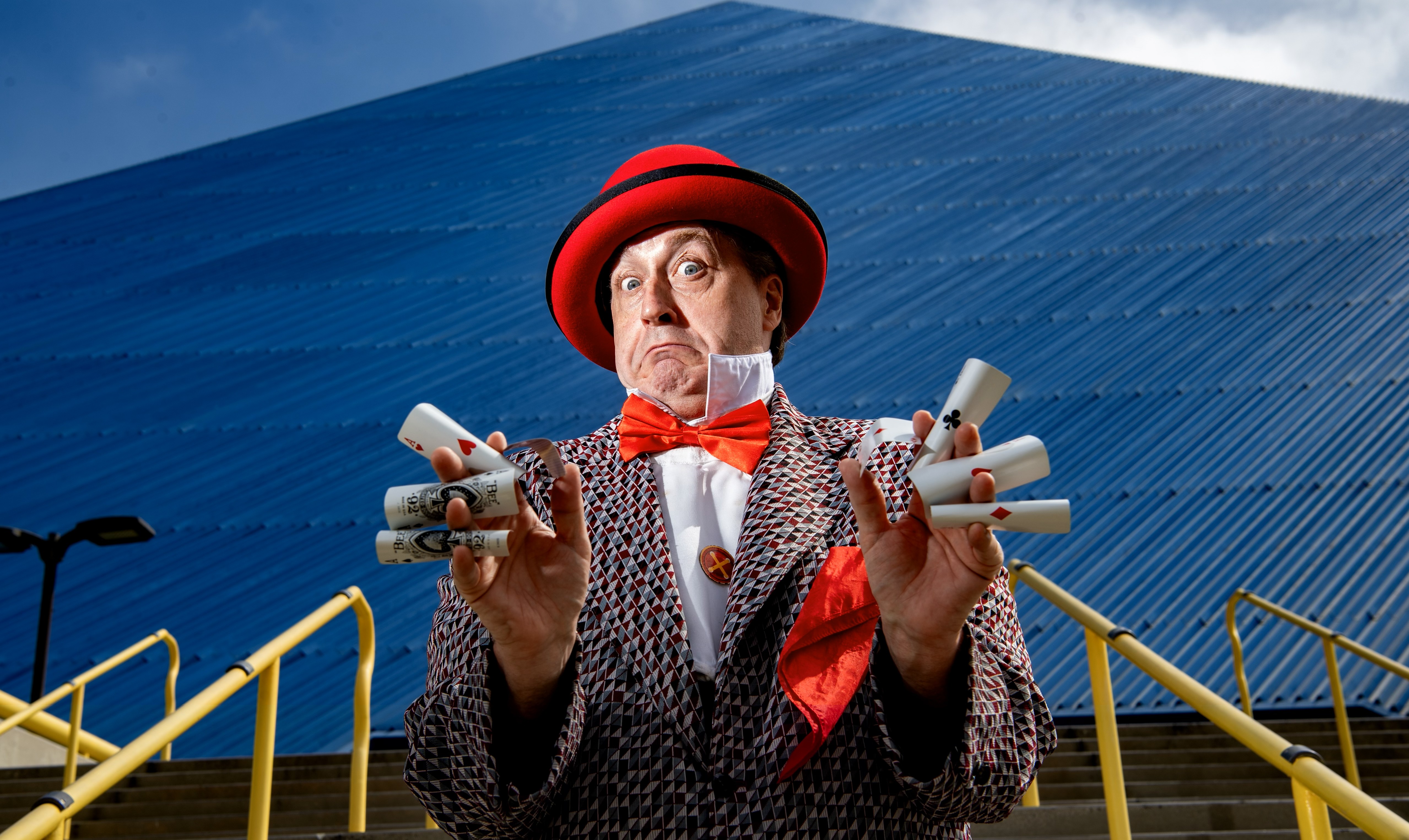 Chris Bange headshot. A man in a polka dot suit and red hat holding playing cards.