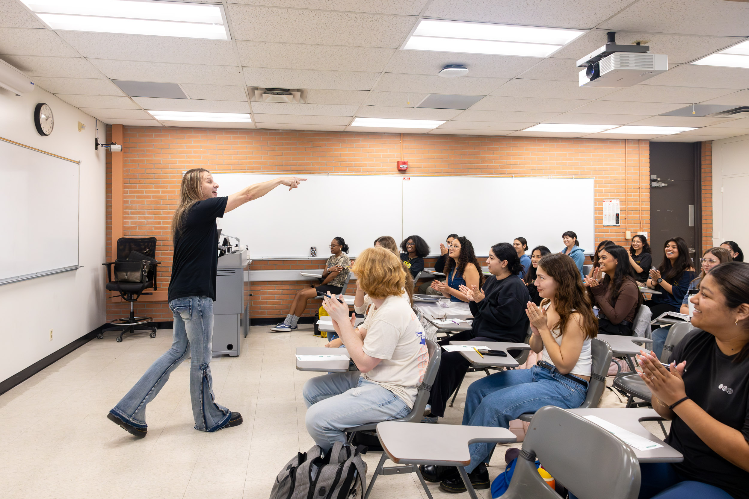 A professor with long hair points toward students as a class applauds in a brightly lit classroom.